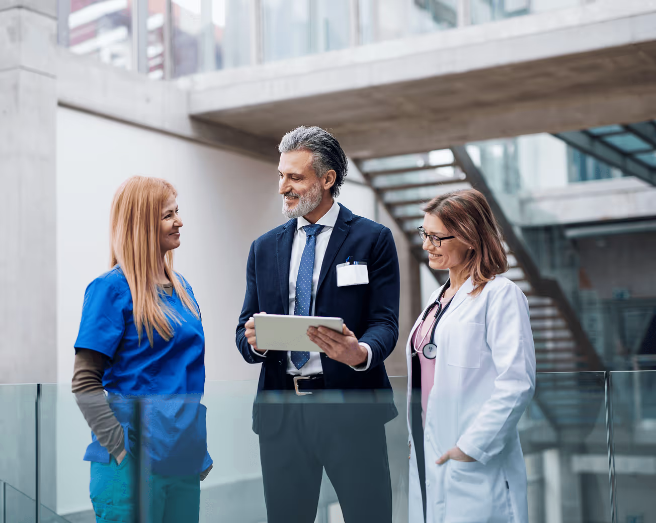 Three healthcare professionals, including a man in a suit with a tablet and two women in medical attire, standing and smiling in a modern building.
